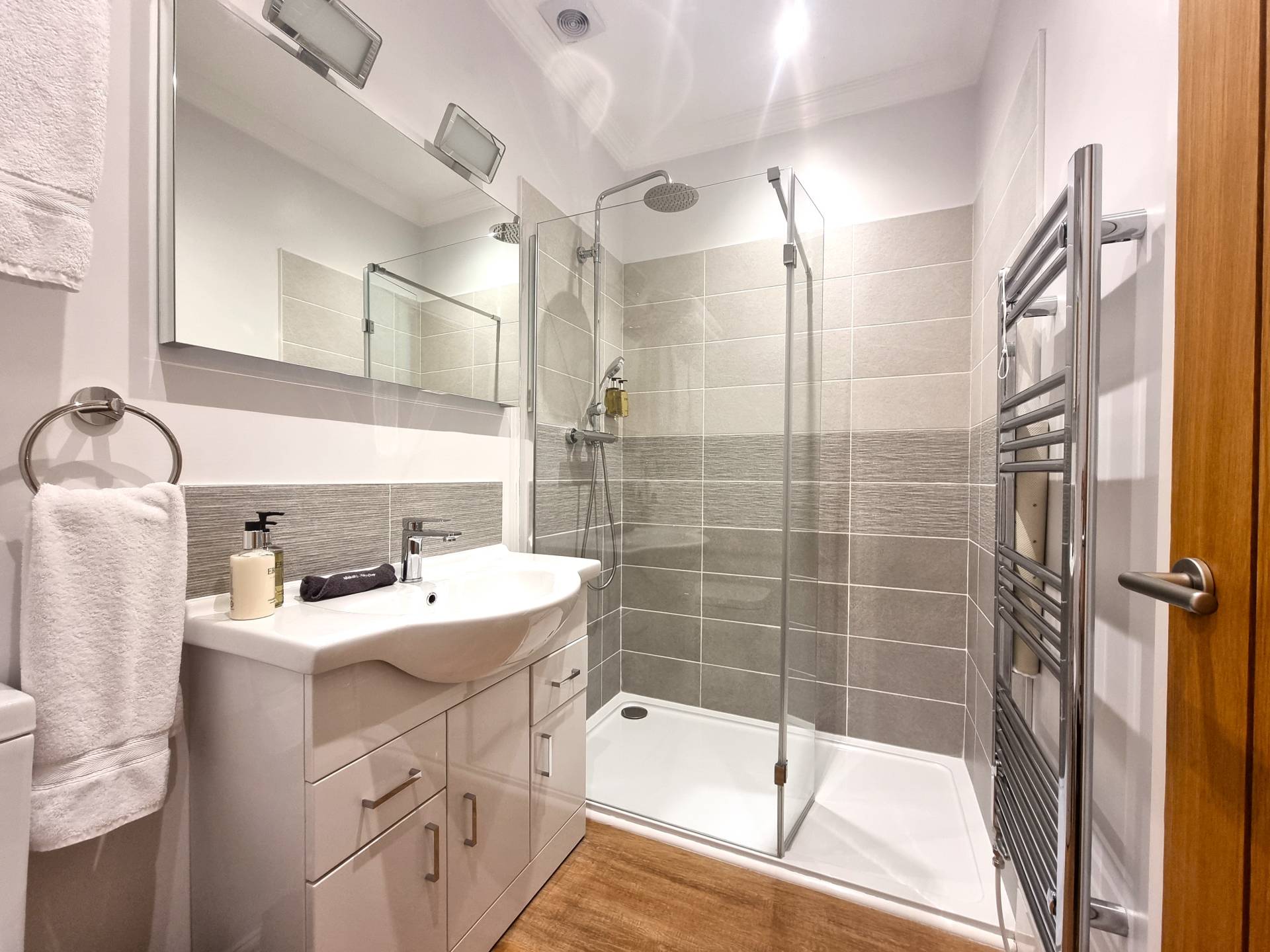 En-suite bathroom in Room 102 at Min y Don Llandudno with walk-in shower, heated mirror with toothbrush and shaver charging point, vanity unit, heated towel rail, and complimentary toiletries including shampoo, moisturiser, hand and body wash, and makeup remover.